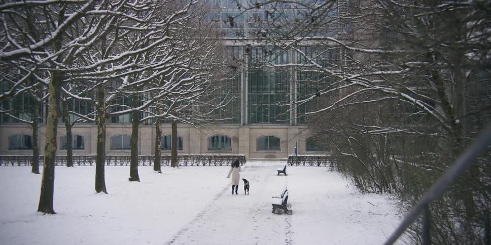 A woman and her dog walk through snow outside of a large building