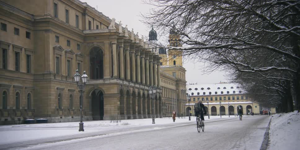 Cyclist and pedestrians move across snow near historic building across street from leafless trees
