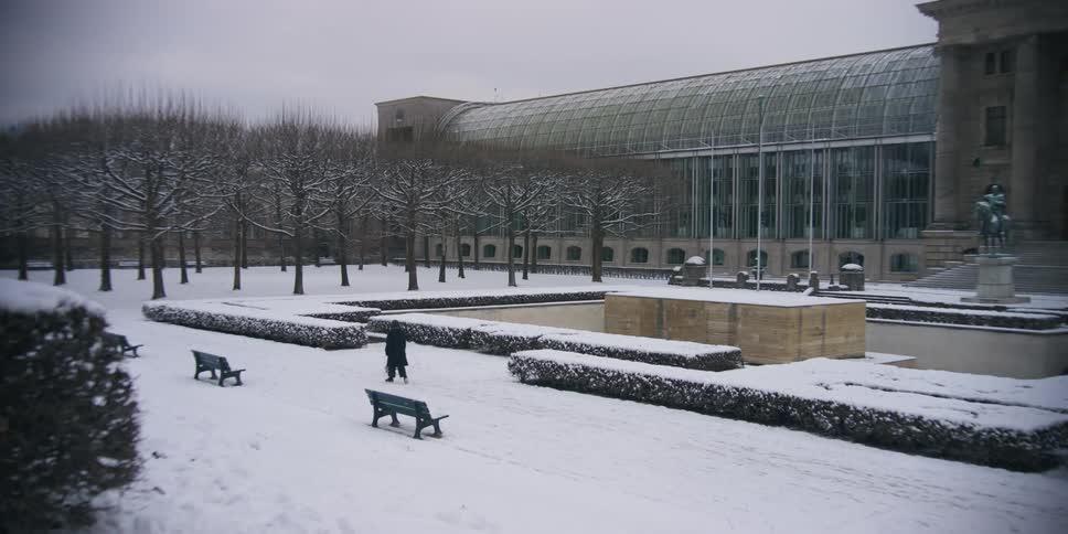 A person is walking a small dog on snowy ground in front of a building