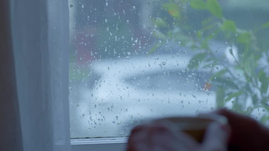 Hands of a person holding a cup that is steaming in front of a window with rain splashes