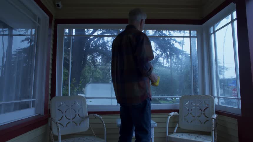 An old man stands in a room and looks out the window