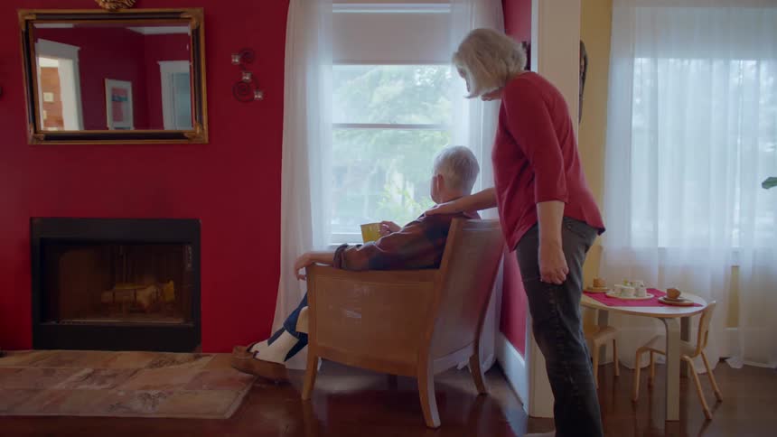 Elderly woman standing massaging the shoulder of her husband who is sitting in a chair holding a cup in front of the window
