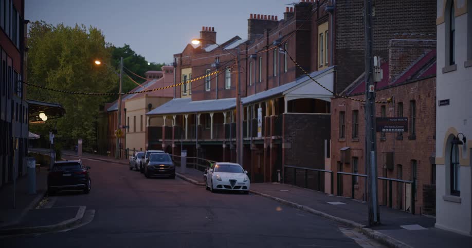 Vehicles sit parked under glowing streetlights on road between brick buildings