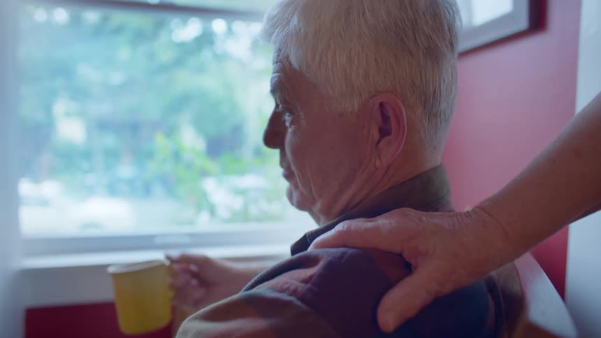 An old man holding a mug is sitting by a window looking outside when an elderly hand comes and massages his shoulder