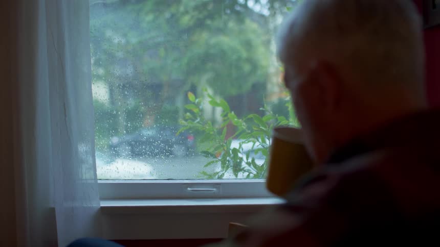 A man looks out a window and sips a drink out of a mug