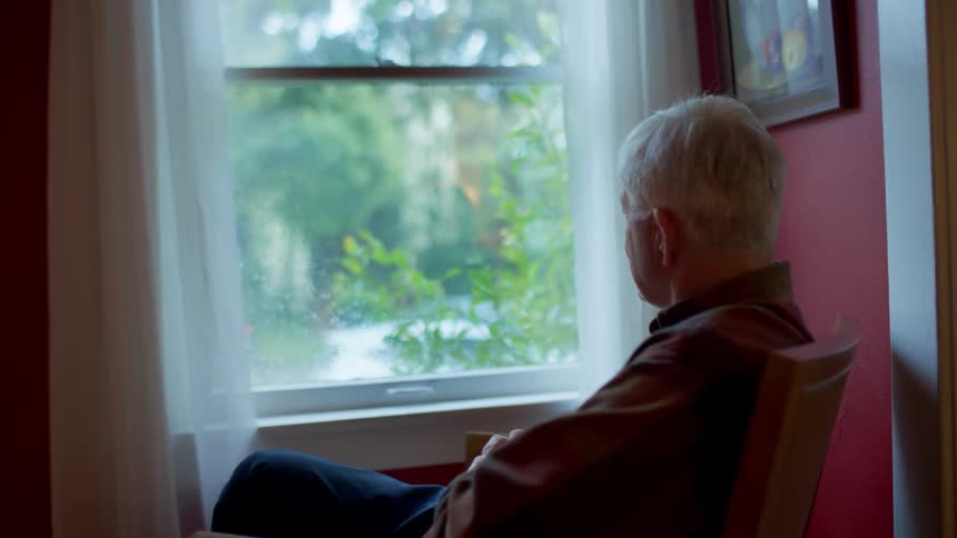 An elderly man sits in front of a window watching the rain