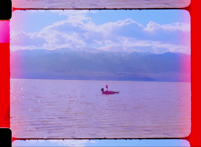 Person lying on a flamingo float in the middle of the sea with mountains around on a blue sky day with clouds