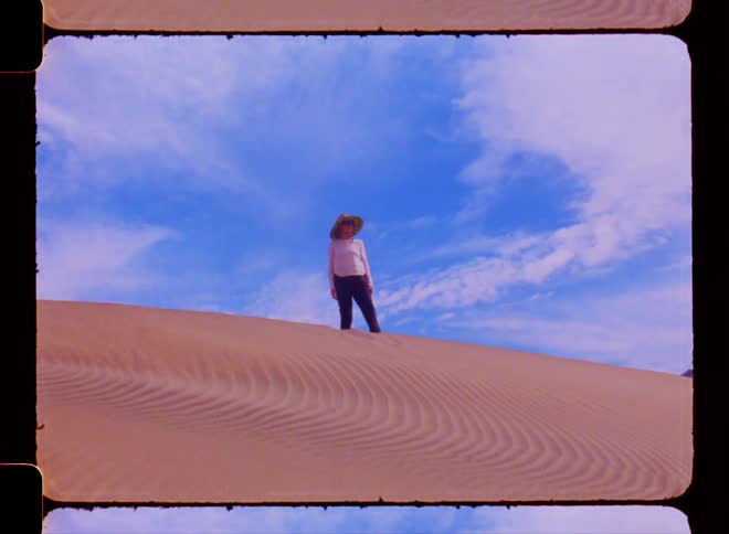 A woman wearing a sun hat stands atop a sand dune under a blue sky with clouds
