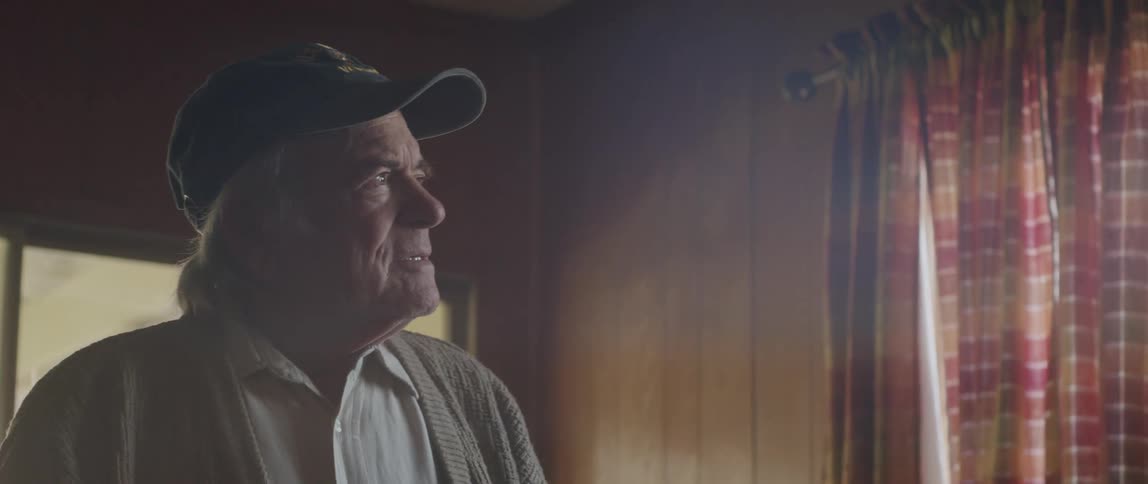 An elderly man walks to a window looks outside and then walks back