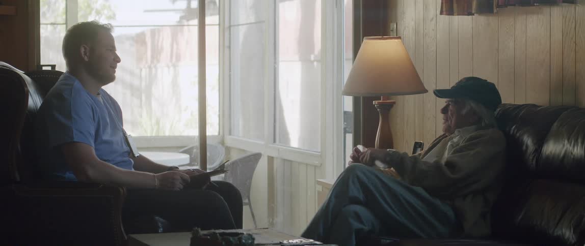 Elderly man sits and talks to male professional care giver wearing scrubs