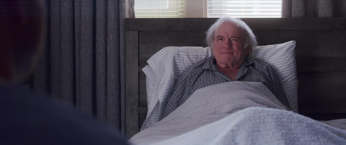 Elderly man lies in bed at home while talking to male caregiver wearing scrubs