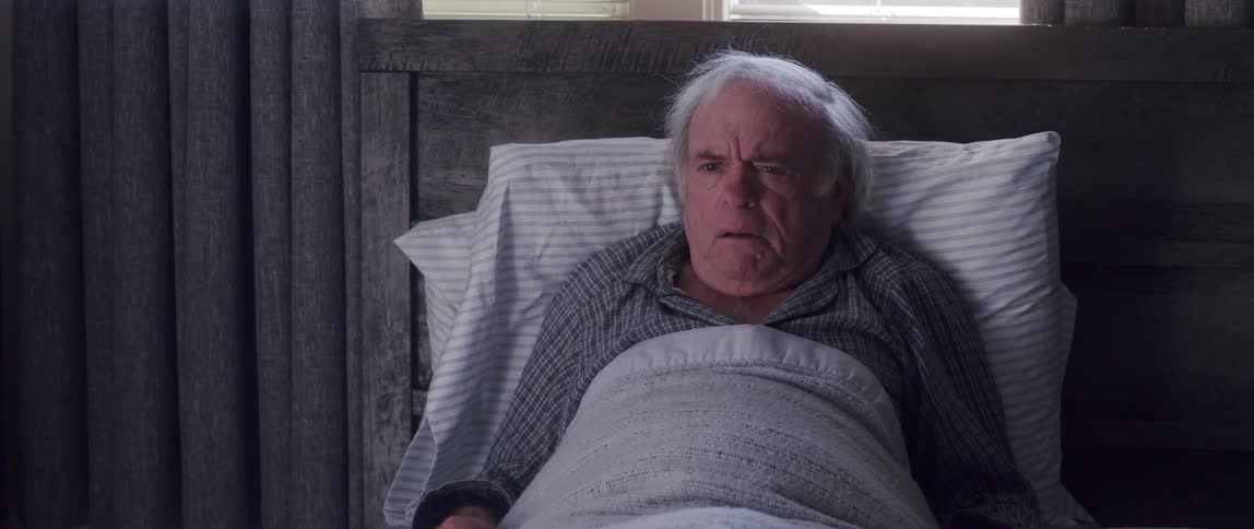 An elderly man is lying on pillows and wearing blankets and has confused expressions