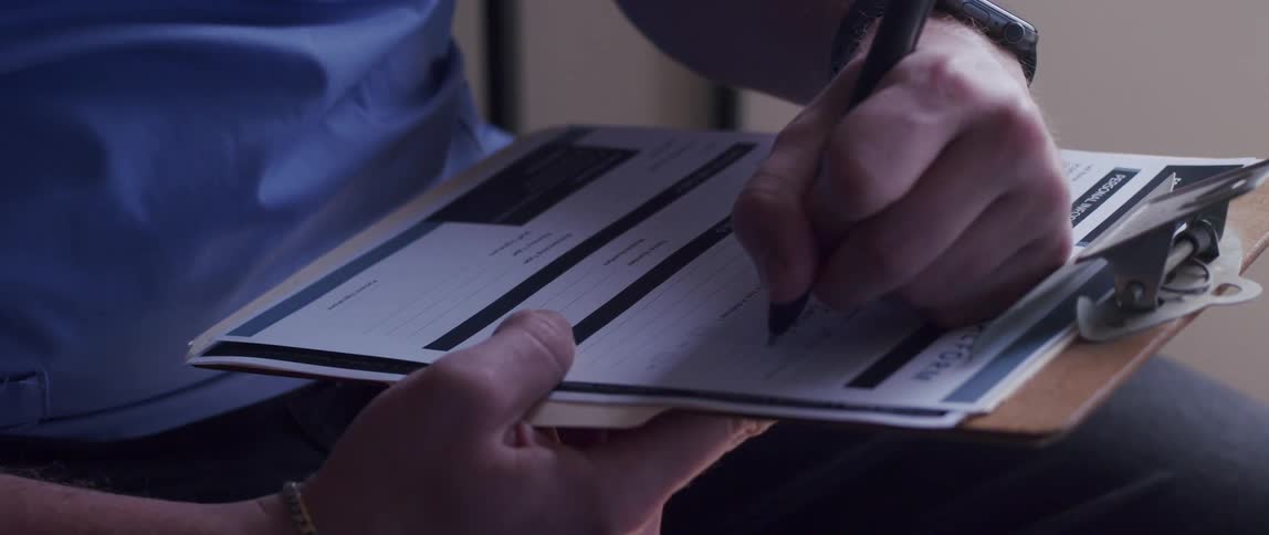 A human hand is writing on paper that is on a clipboard using a pen