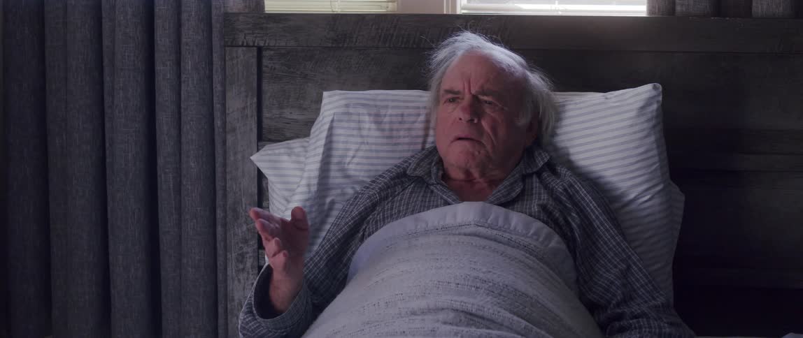 An elderly man is lying down with several pillows on his back and starts talking to someone who passes in front of him