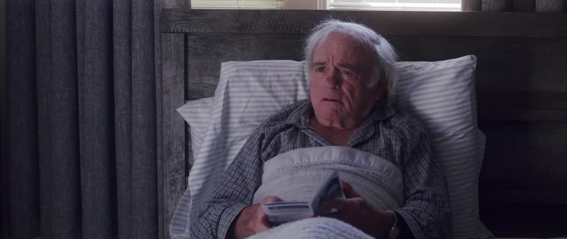 Elderly man sitting on a bed taking a pamphlet that a person hands him with a sad facial expression
