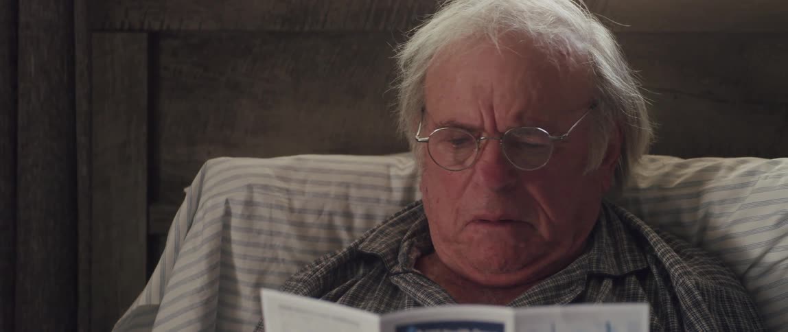 Elderly man sitting on his bed reading a pamphlet and then removing his prescription glasses