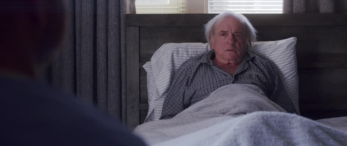 An old man sitting up on a bed looks nervous and scared as a doctor talks to him