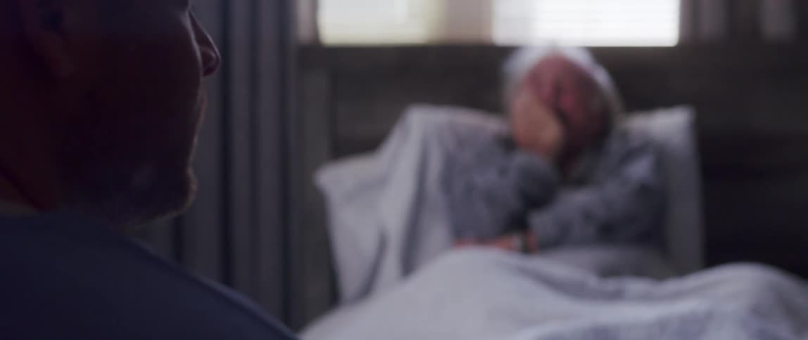 An elderly man lying down is crying and passing his hand over his face while there is a person in front of him watching and then looking to the side