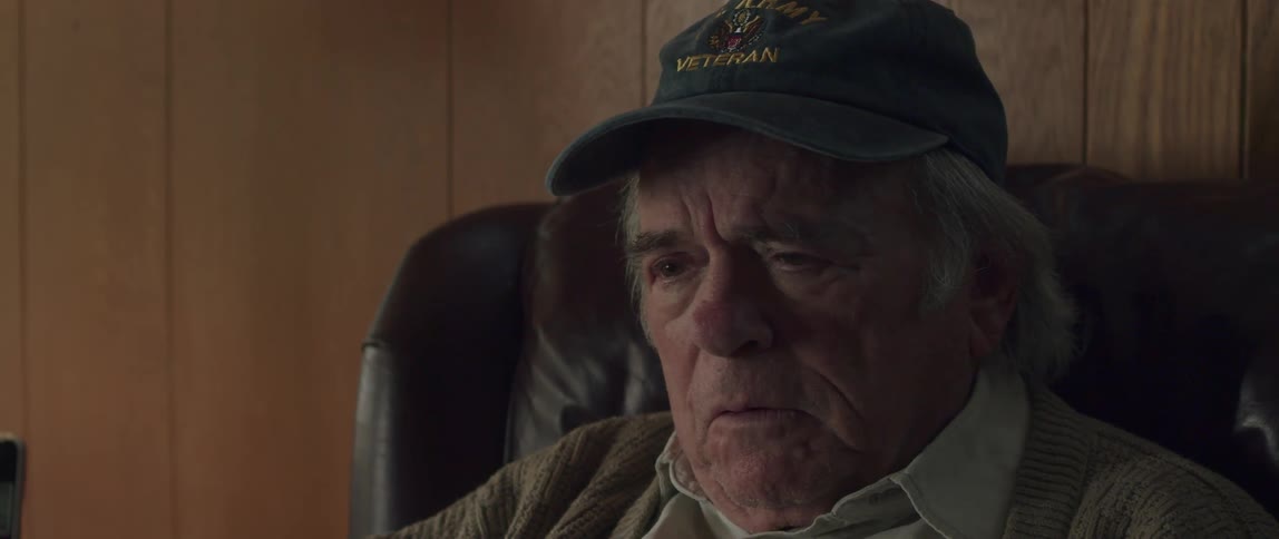 Elderly man wearing a baseball cap sitting on a chair at home talking