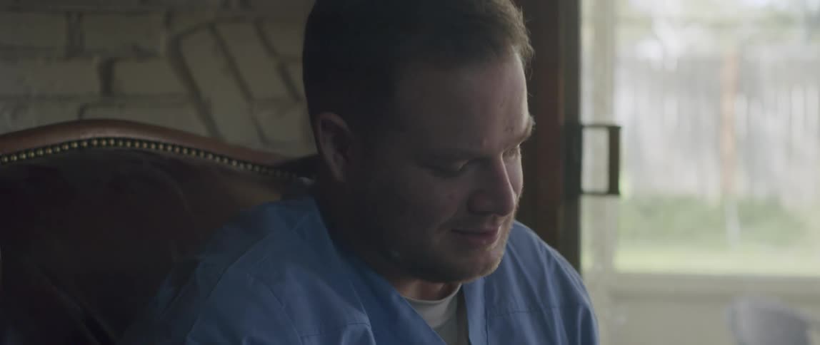A doctor sitting on a chair talks and scratches his nose while holding a pen