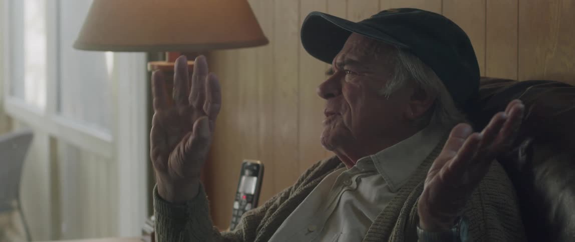 An elderly man in a cap is sitting and talking and raises his hands