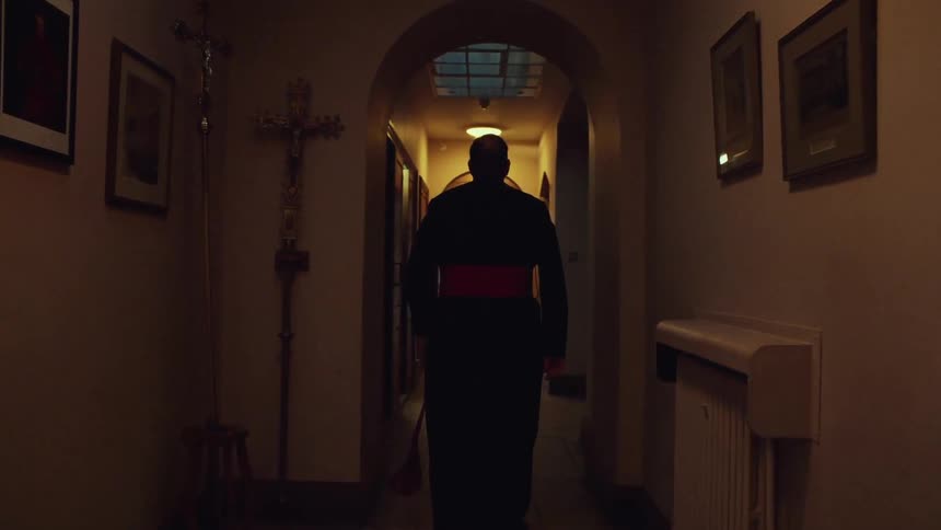 Priest in a black robe walks down the hallway of a church