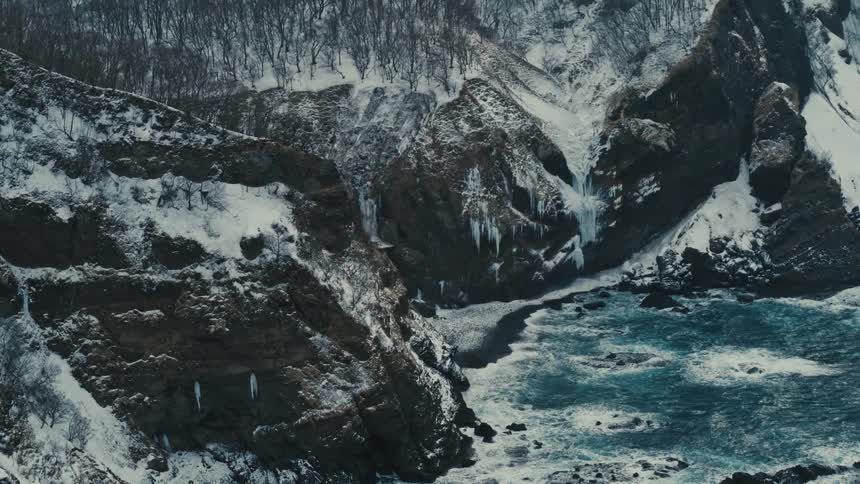 Waves roll in and splash against rocks along the snow covered cliff on the coast