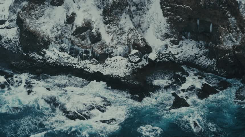 Aerial in light snow fall of waves crashing onto snow covered arctic rockshore