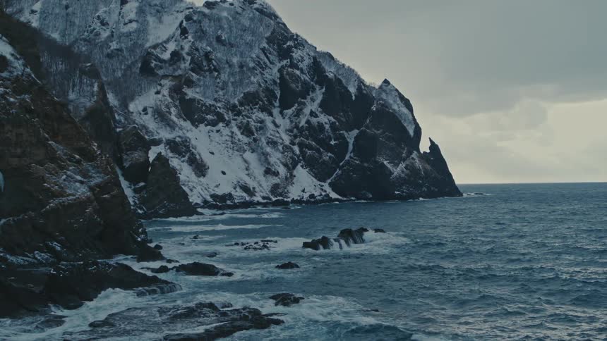 Waves crash upon rocks in the ocean at the base of snowcovered mountains