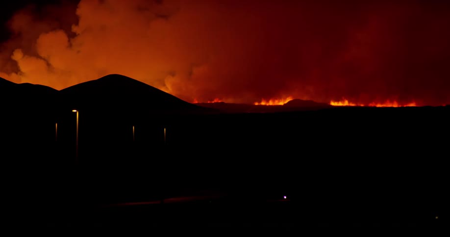 Glowing red smoke fills the sky as flames rise in the distance and traffic passes on the road