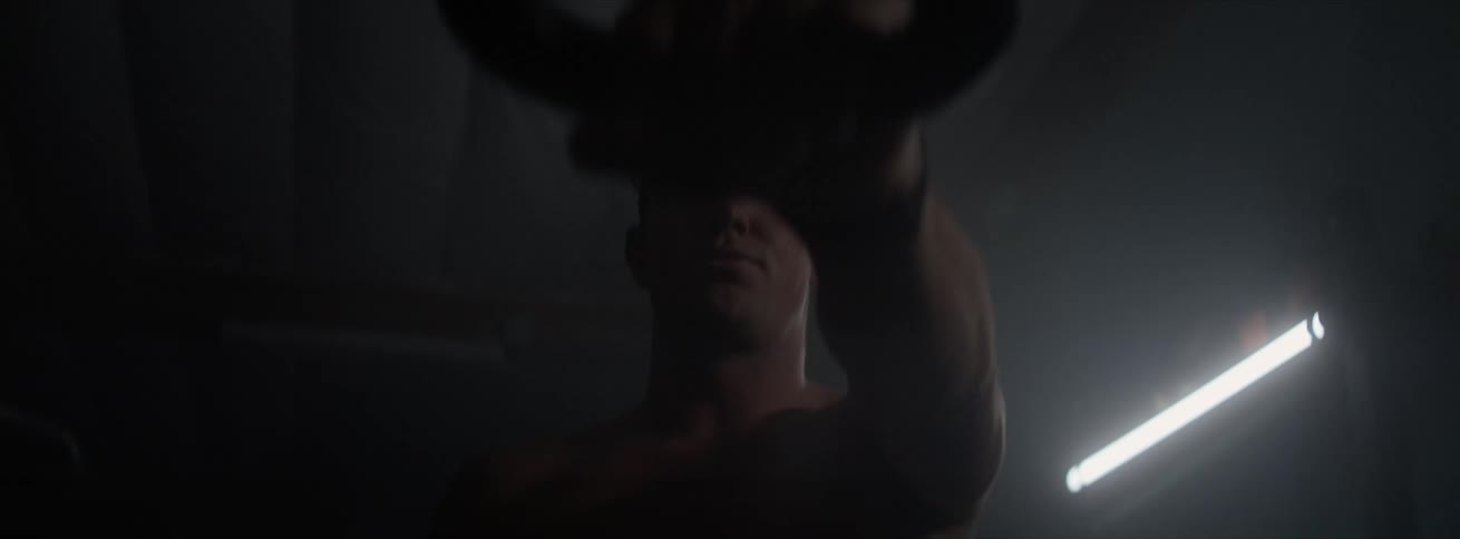 Man without a shirt lifting a weight with one of his arms extended in a lowlight environment