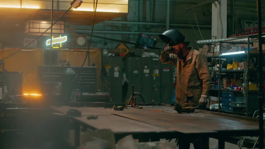 A metal worker with his safety mask turned up cleans the sweat off his forehead with his sleeve