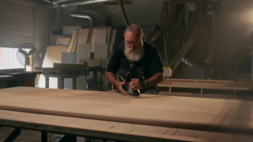 Bearded woodworker sands a long board of wood in a workshop