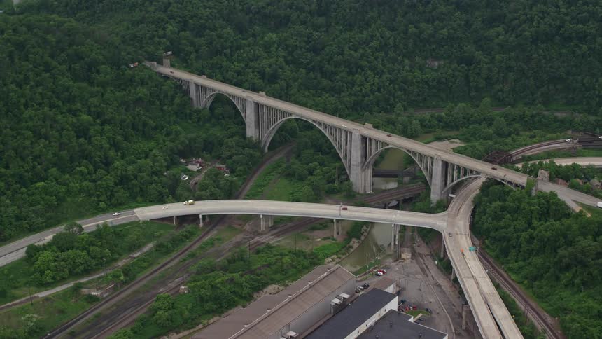 Aerial view of a large bridge over highways and river with vehicle traffic amid forested hills and mountains