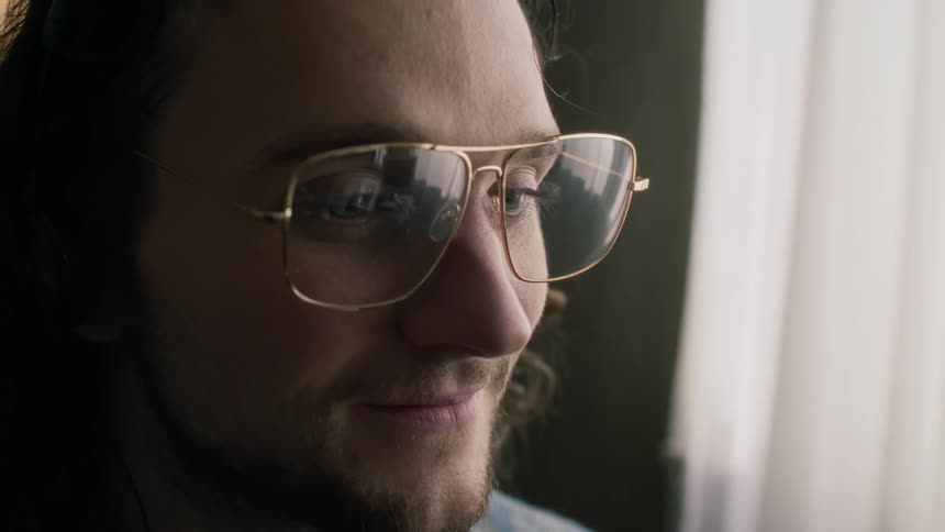 Close up of the face of a young bearded man with long hair and glasses