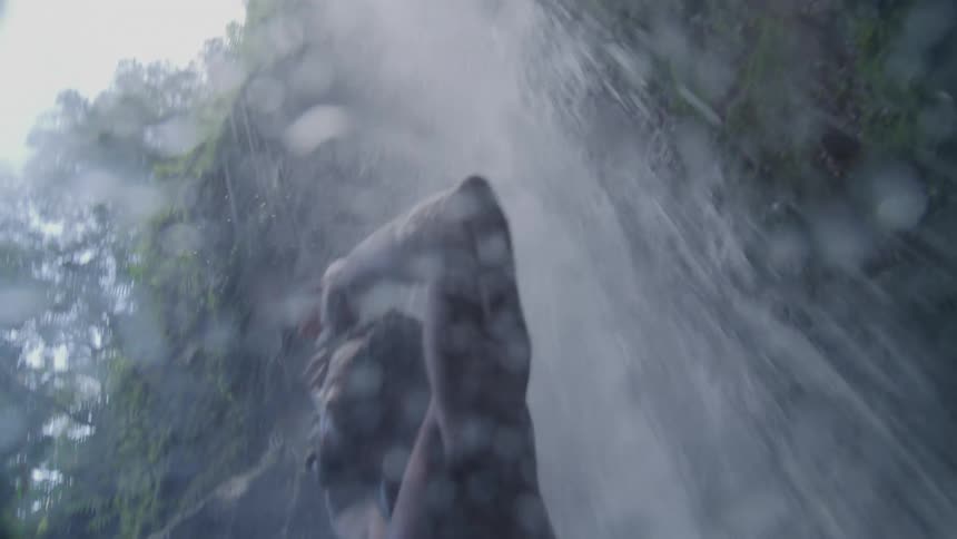 Man in just shorts and no tshirt under a waterfall with his arms up