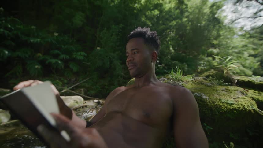 Man without tshirts in shorts lying on rocks next to a river reading a book