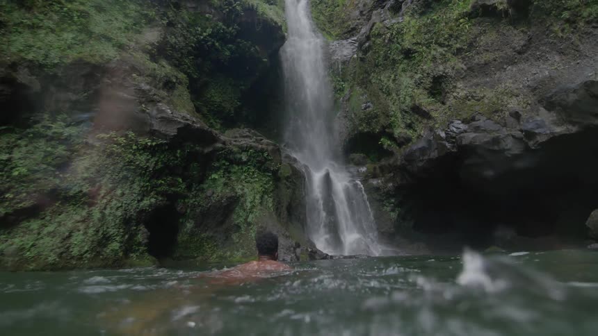 Back view of a man swimming in a river towards a cascade waterfall