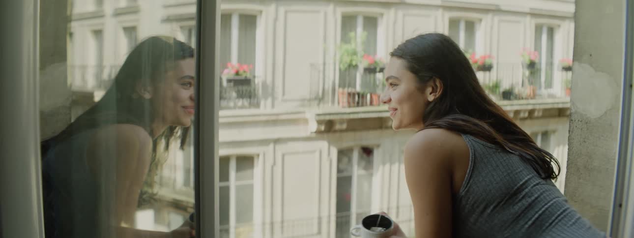 A young woman holding a tea cup leans out of a window to look at the view of the city smiling