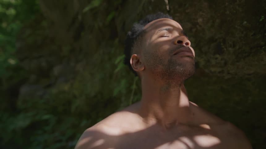 Portrait of a man closing his eyes and taking a deep breath while standing next to a rock in a natural environment with a sun flare