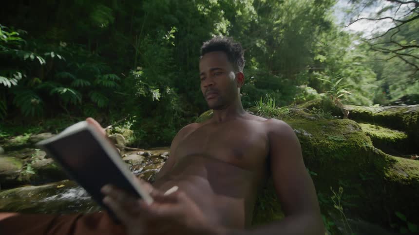Man in a swimsuit reads a book next to a stream in the forest