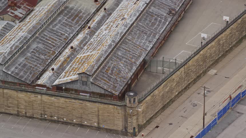 A prison sits inside a brick wall fence with a large parking lot outside of it
