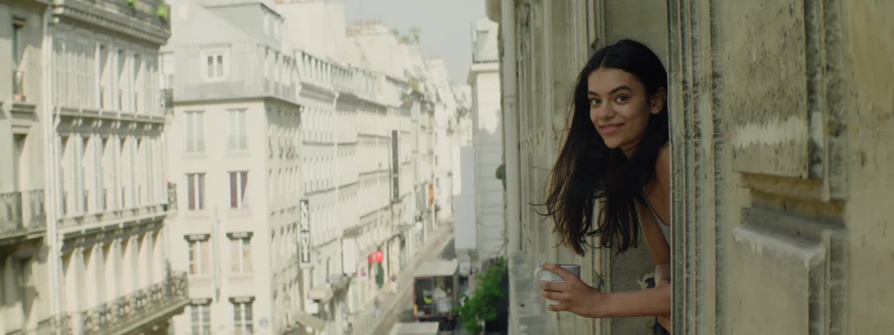 A young girl leaning out of a window holding a cup looking at a city street with classical architecture