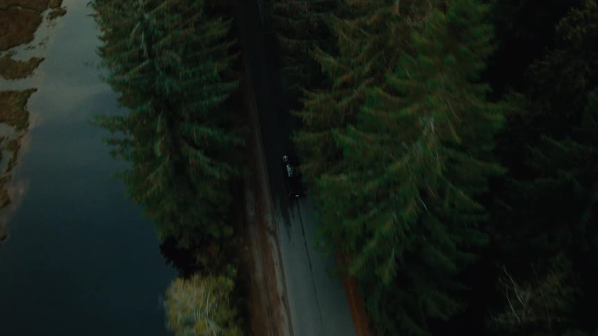 Aerial view of a black car driving on a road next a river in the mountains