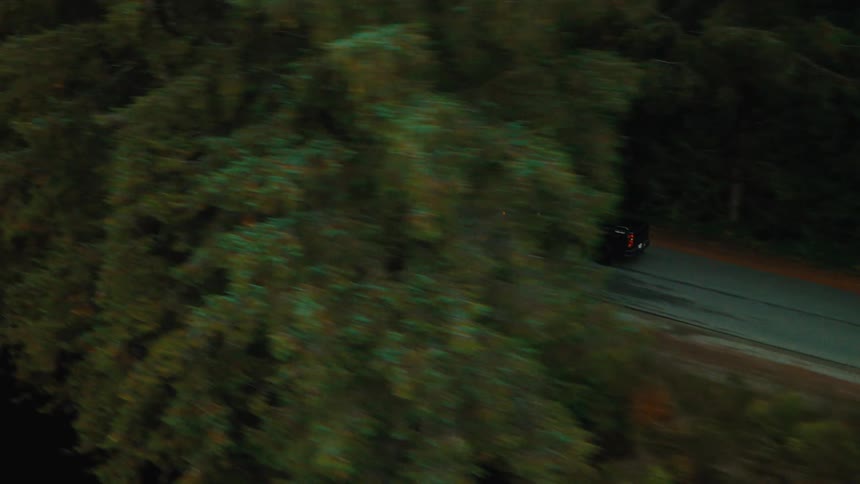 Aerial view of a black pickup truck moving along a narrow street next to a lake and forest