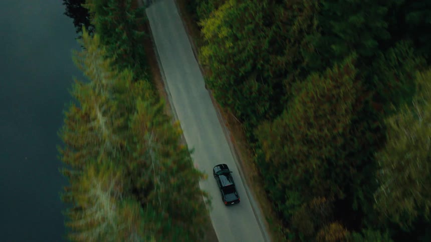 A black car driving on a road surrounded by pine trees and a lake reflecting the trees