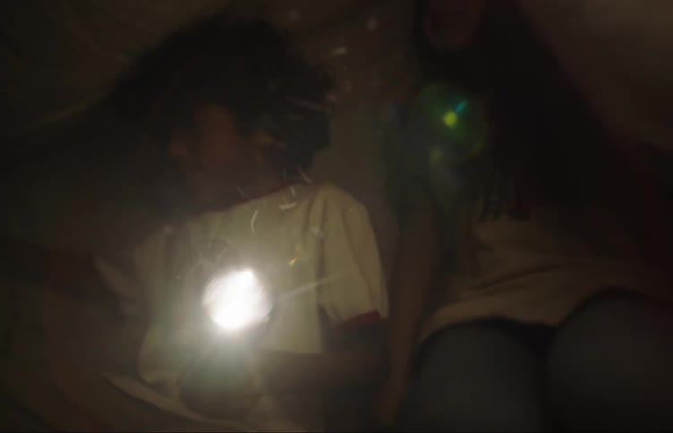 A boy and a girl are happy and jumping holding a flashlight and there is a sheet on top of them