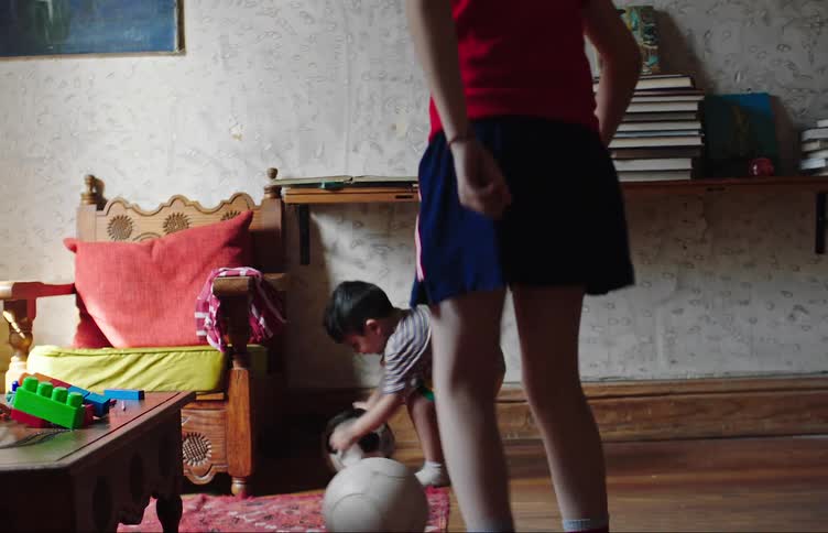 A little boy kicks a soccer ball to a bigger boy in a room with lots of toys and they both start playing with soccer balls