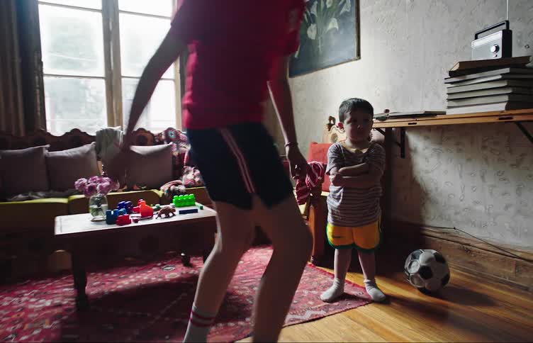 Boy dribbles a soccer ball around the living room as his pouty younger brother looks on