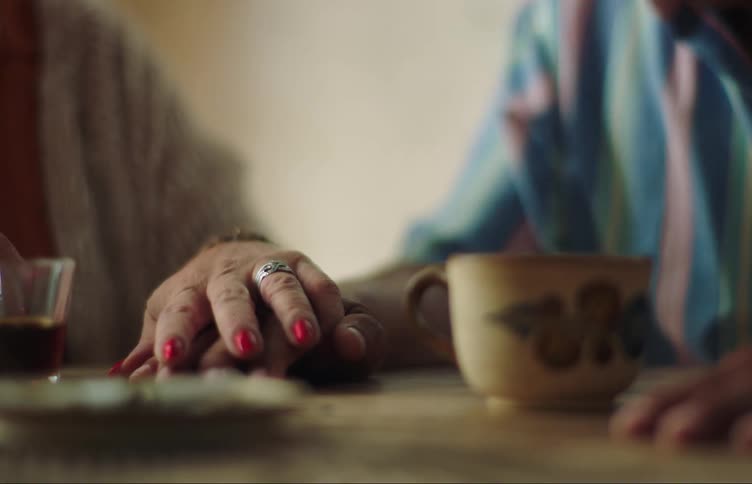 A man and a woman are sitting at a table having coffee and tea when the man angrily hits his hand on the table and the woman gently puts her hand on his to calm him down